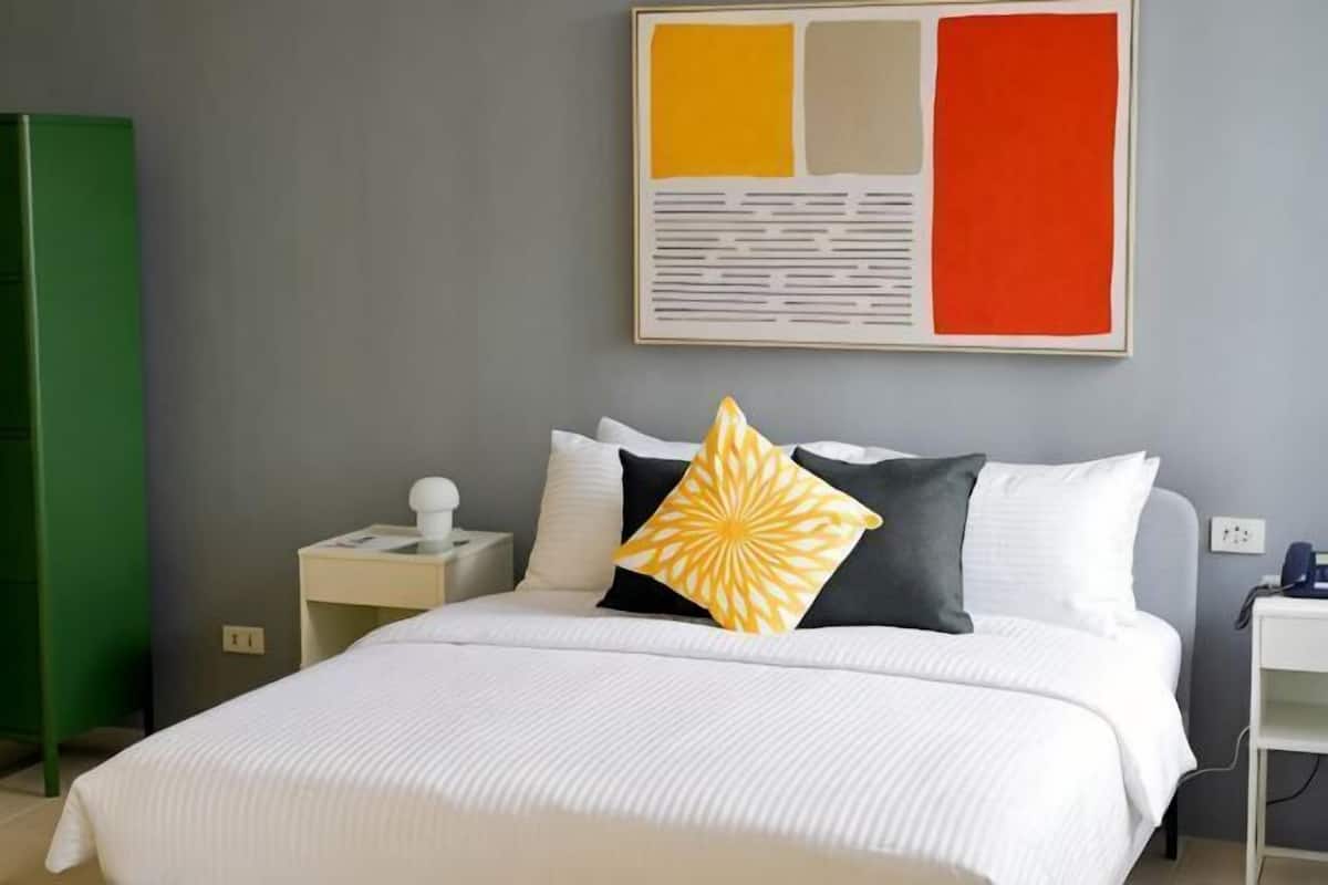 A hotel room with a bed, a nightstand, a green wardrobe, and a wall with a large abstract painting.