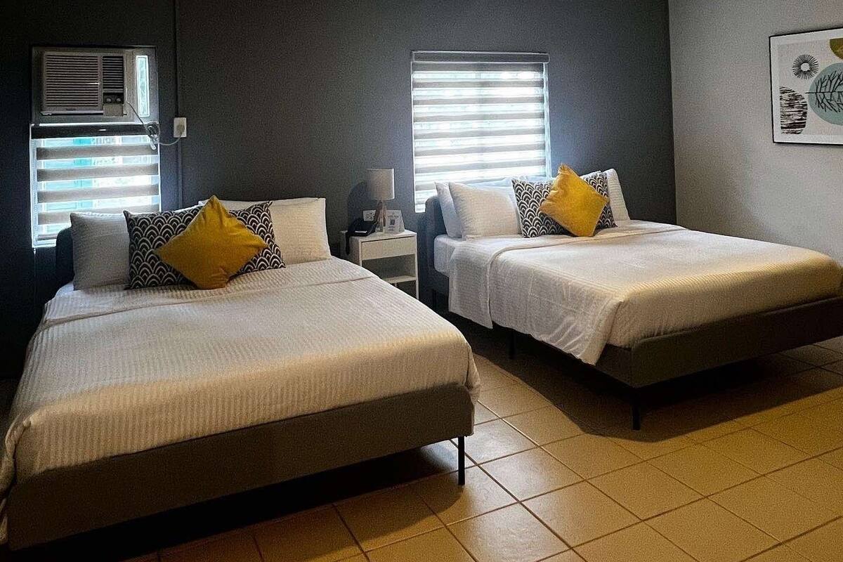 A hotel room with two beds, a small bedside table with a lamp, and a wall-mounted air conditioning unit.