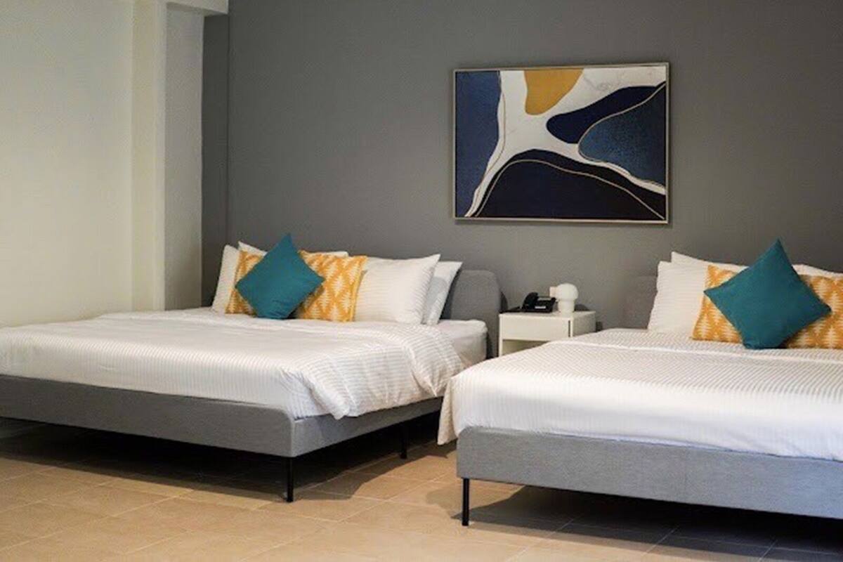 A hotel room with two beds, each with white bedding and teal pillows. There is a gray wall with a large abstract painting above the beds.