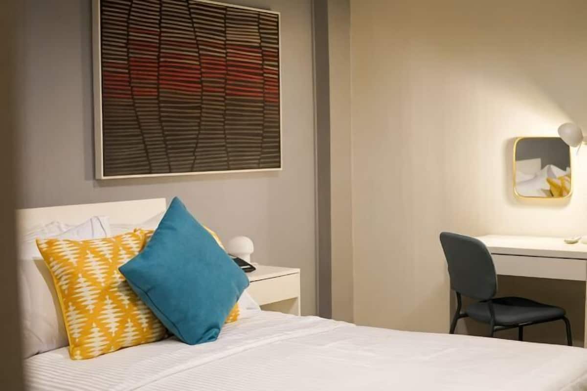 A hotel room with a bed, a desk, a chair, a wall-mounted artwork, and a window with blinds.