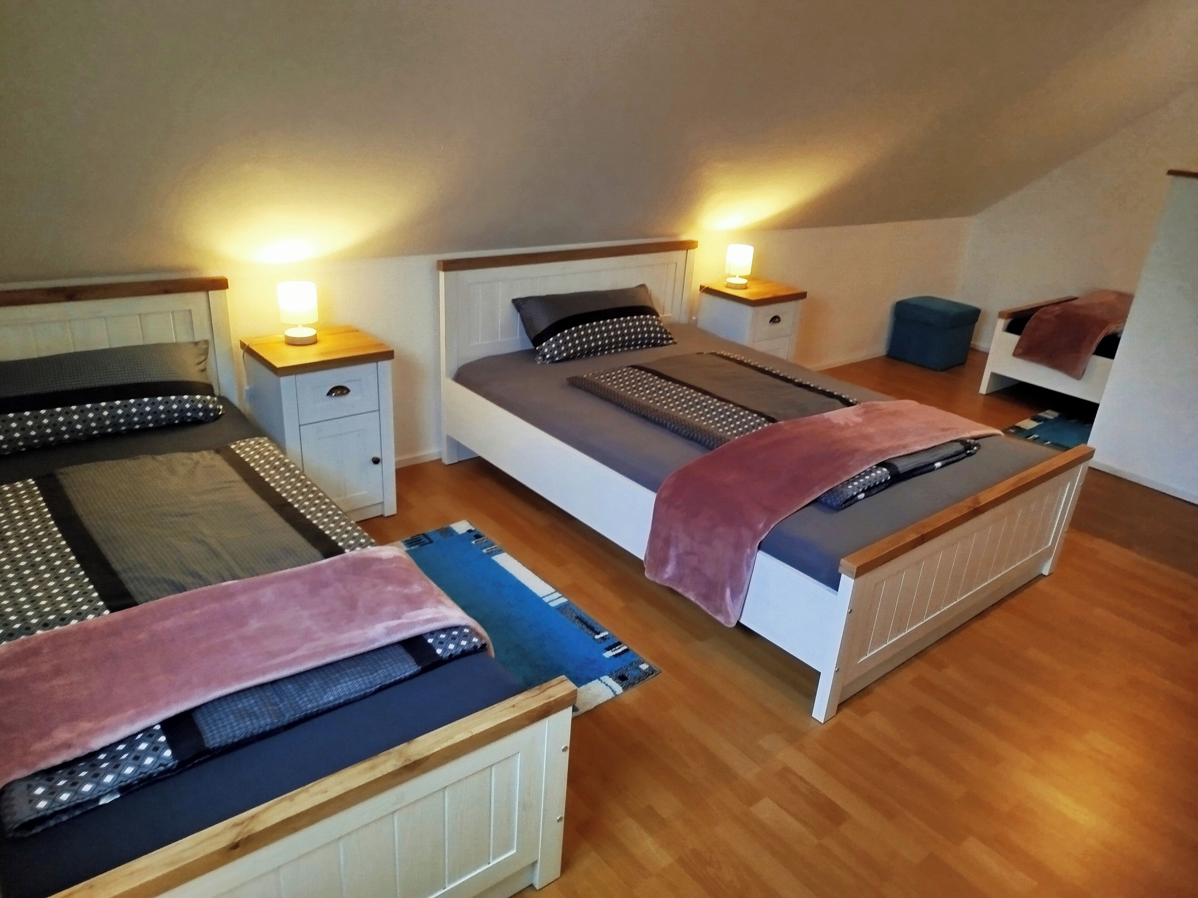 Ein Zimmer mit zwei Betten, jeweils mit einem Nachttisch und einer Lampe. Der Raum hat Holzboden und eine schräge Decke.