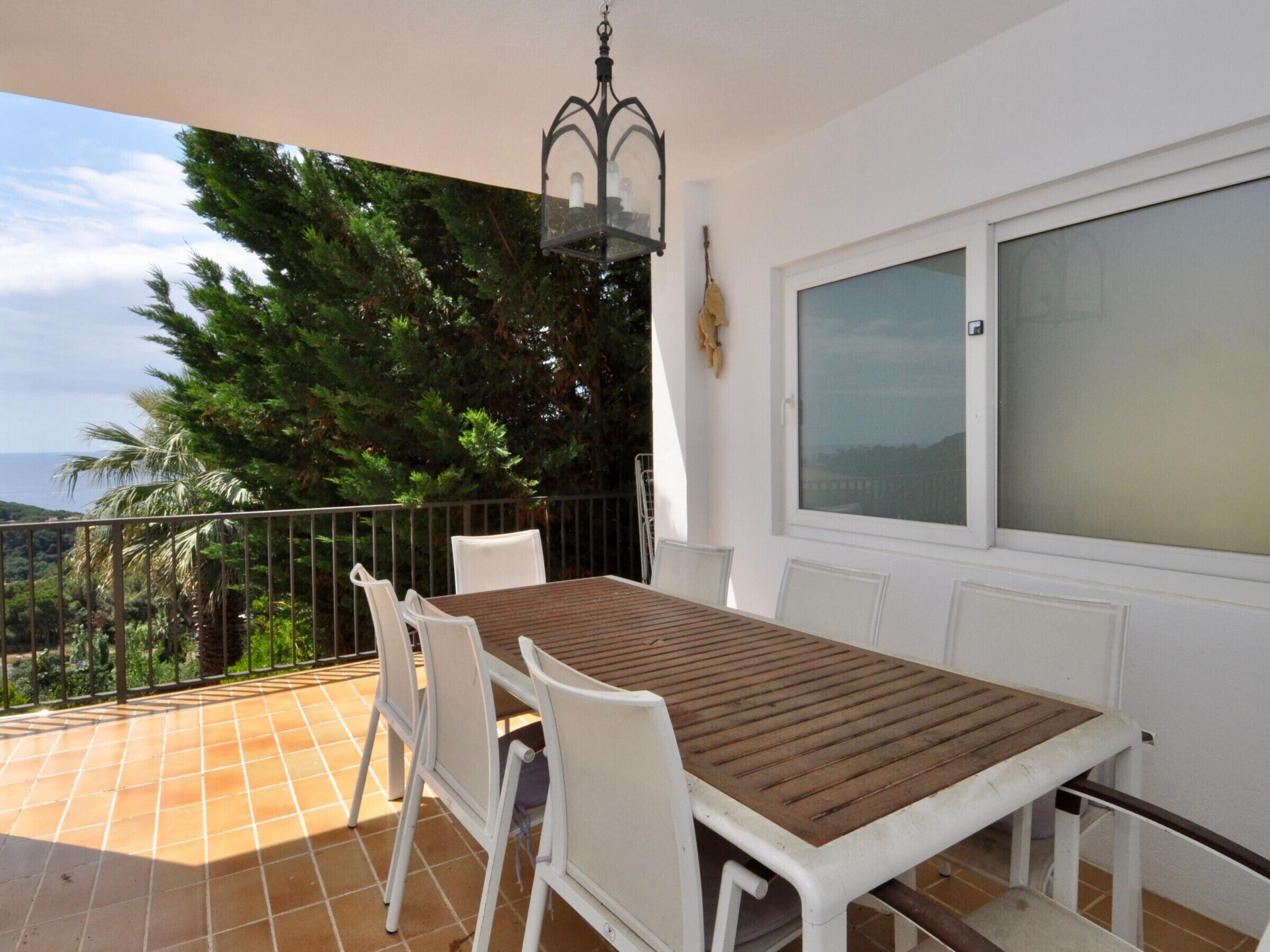 A balcony with a table and chairs, a window, and a view of trees and the sea.