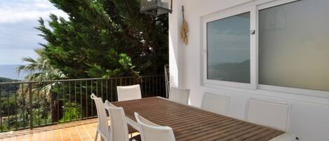 A balcony with a table and chairs, a window, and a view of trees and the sea.