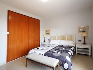 A bedroom with a bed, a bench, a wardrobe, two bedside tables with lamps, and two framed pictures on the wall.