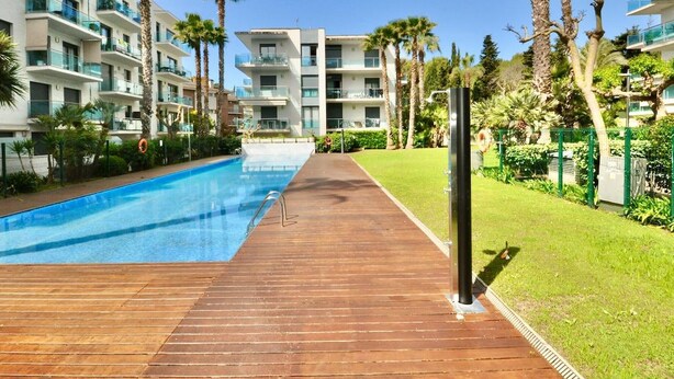 Un área de piscina con muelle de madera, rodeada de edificios de varios pisos con balcones, palmeras y cielo despejado.
