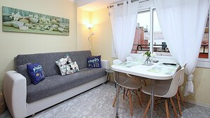 A living area with a grey sofa, two patterned cushions, a white table with chairs, a window with white curtains, a floor lamp, and a painting on the wall.