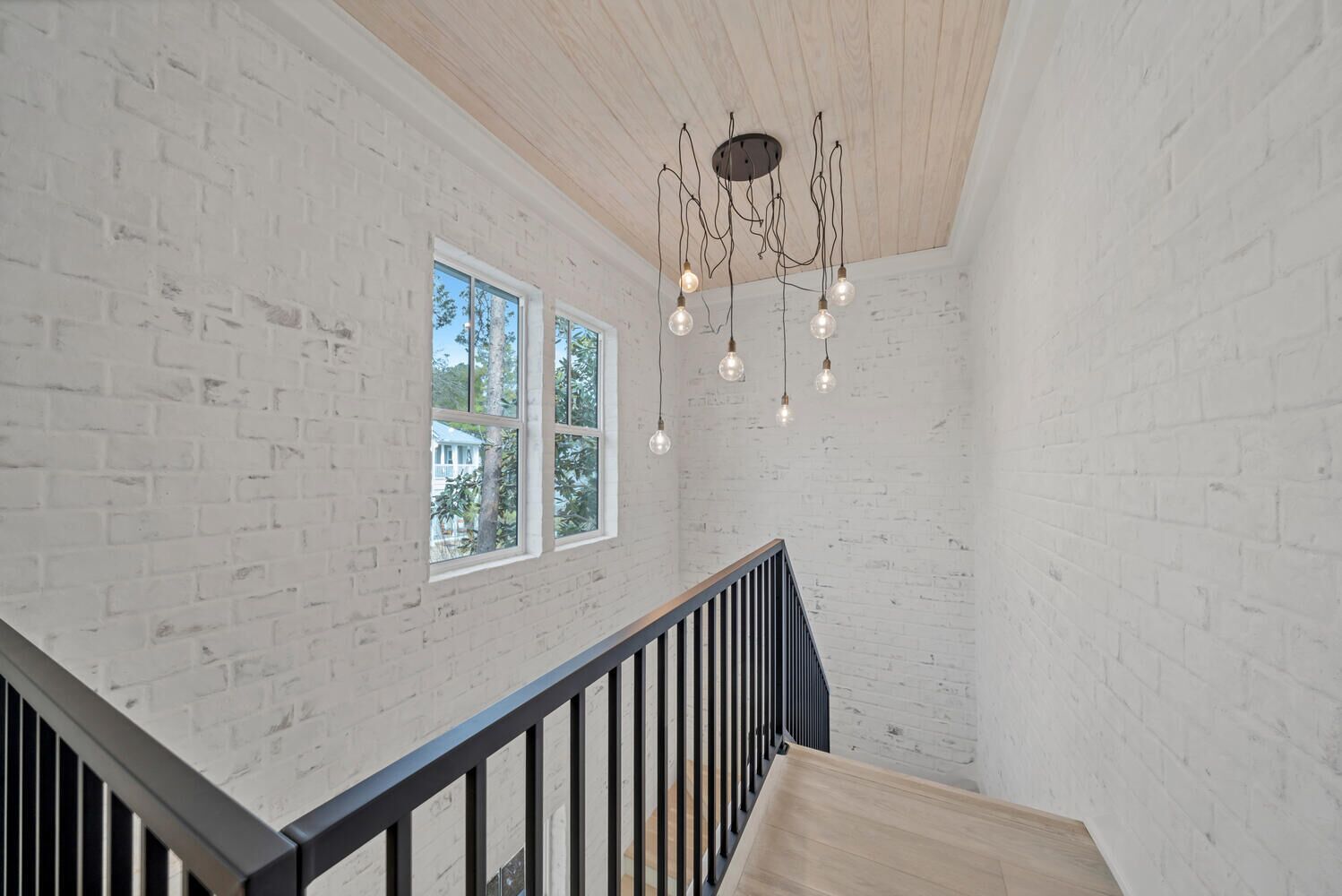 A room with white brick walls, a wooden ceiling, and a black railing. There are two windows with a view of trees and a house.