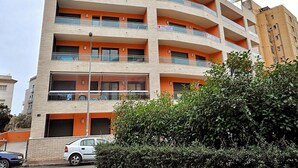 A multi-story building with balconies, a car parked in front, and a hedge in the foreground.