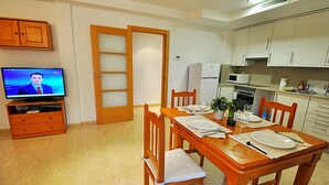 A dining area with a wooden table set for four, a television on a wooden stand, and a kitchen with white cabinets and appliances.