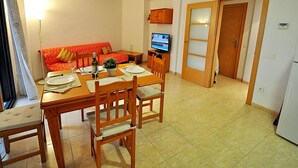 A dining area with a wooden table set for two, a sofa with a red cover, and a television on a stand.