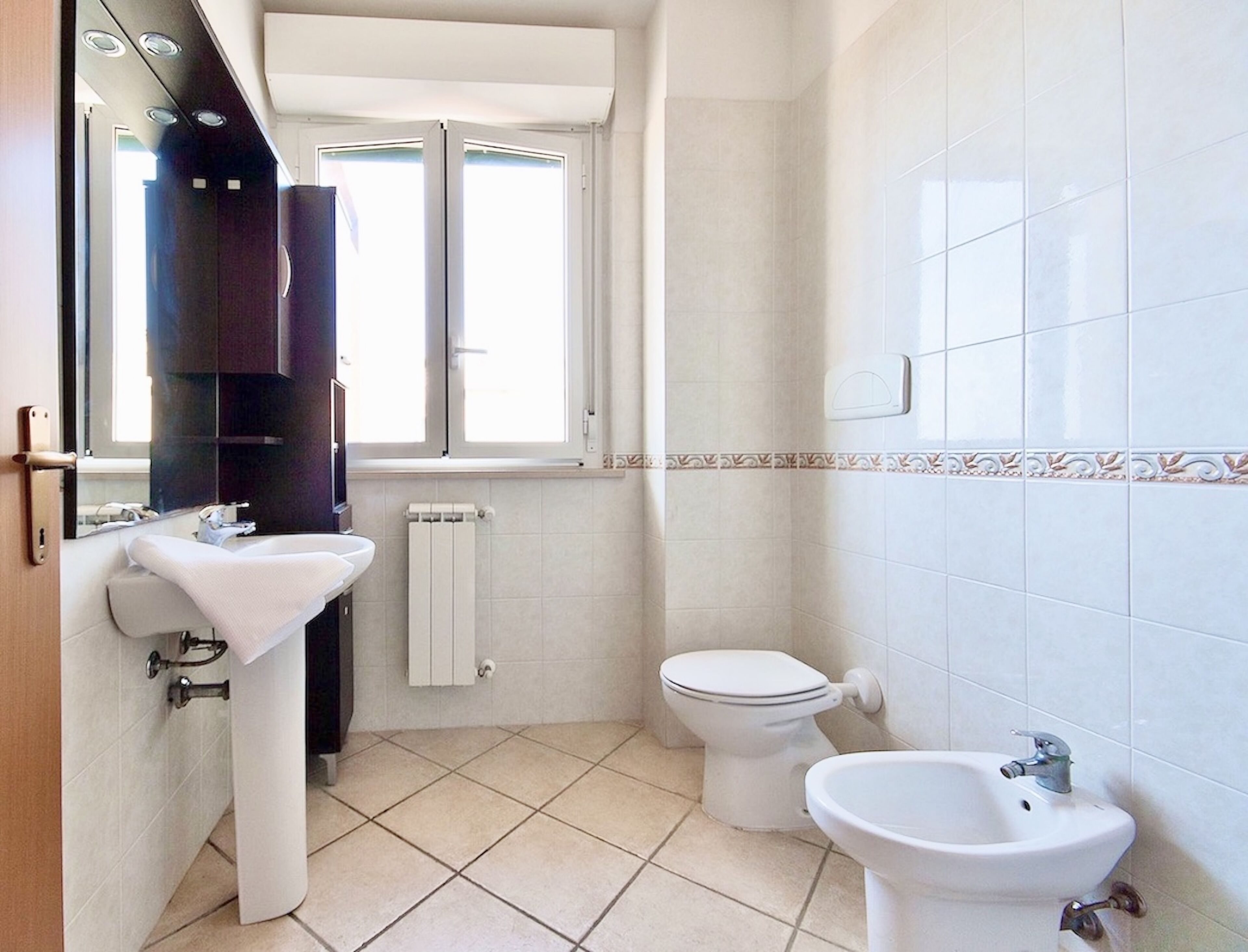Une salle de bain avec un lavabo blanc, des toilettes et un bidet. Il y a une fenêtre au-dessus des toilettes et un radiateur en dessous.