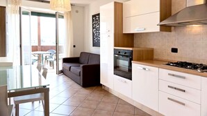 A modern kitchen with white cabinets, a gas stove, and a built-in oven. There is a glass dining table with chairs, a sofa, and a view of the outdoors through a sliding glass door.