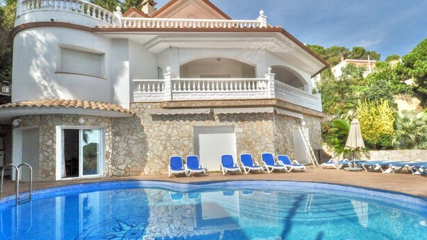 A two-story building with a swimming pool, stone walls, and a balcony with blue lounge chairs.