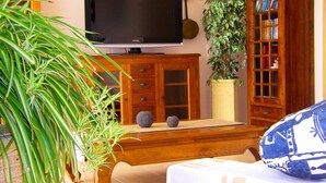 Een woonkamer met een televisie op een houten standaard, een kamerplant, een boekenkast en een biljarttafel.