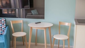 A room with a light blue wall, a round wooden table with two chairs, a TV mounted above the table, and a blue sofa with a geometric-patterned pillow. - Comfortable Studio | Parking, Balcony, Close to the Beach (Miedzyzdroje)