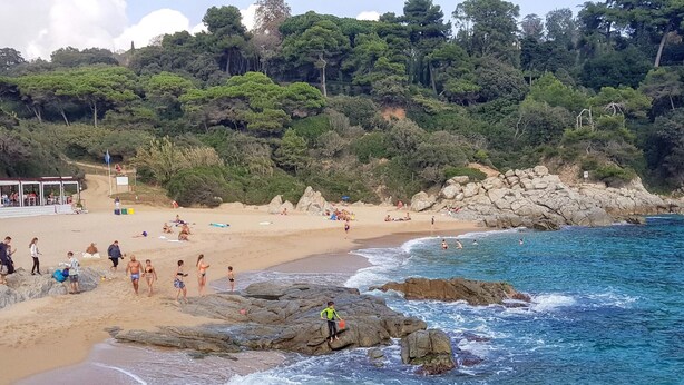 Una spiaggia con persone che nuotano e passeggiano lungo la riva, circondata da rocciame e fitta vegetazione.