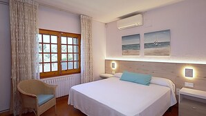 A bedroom with a large bed, a wicker chair, a window with curtains, and wall-mounted artwork. - 6-Bedroom Villa with Private Pool in Lloret de Mar (Lloret de Mar)