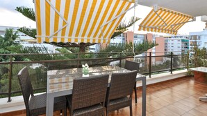 A balcony with wicker chairs and a glass table, yellow and white striped umbrellas, and a view of buildings and greenery. - Cozy Apartment with Pool, 150m from Beach - Lloret de Mar (Lloret de Mar)
