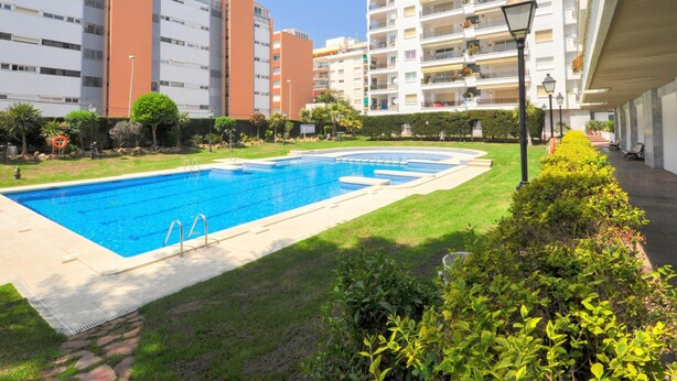 A swimming pool surrounded by a well-maintained lawn, with modern apartment buildings in the background. - Cozy Apartment with Pool, 150m from Beach - Lloret de Mar (Lloret de Mar)