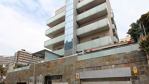 A multi-story building with a modern design, featuring a mix of glass and metal balconies, a visible entrance, and a closed garage door. - Cozy Apartment with A/C, 150m from Lloret de Mar Beach (Lloret de Mar)