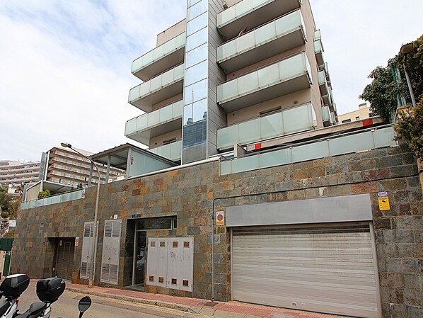 A multi-story building with a modern design, featuring a mix of glass and metal balconies, a visible entrance, and a closed garage door.