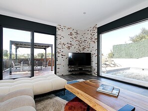 Ein modernes Wohnzimmer mit großem Fernseher, einer Couch, einem Holztisch und Blick auf einen Pool und einen Außenbereich.