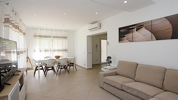 Uma sala de estar moderna com um sofá bege, uma área de jantar com mesa e cadeiras, e uma cozinha com fogão e armários.