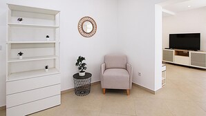 Uma sala de estar moderna com uma estante branca, um espelho redondo, uma pequena planta em vaso, uma poltrona rosa e uma televisão sobre um móvel.