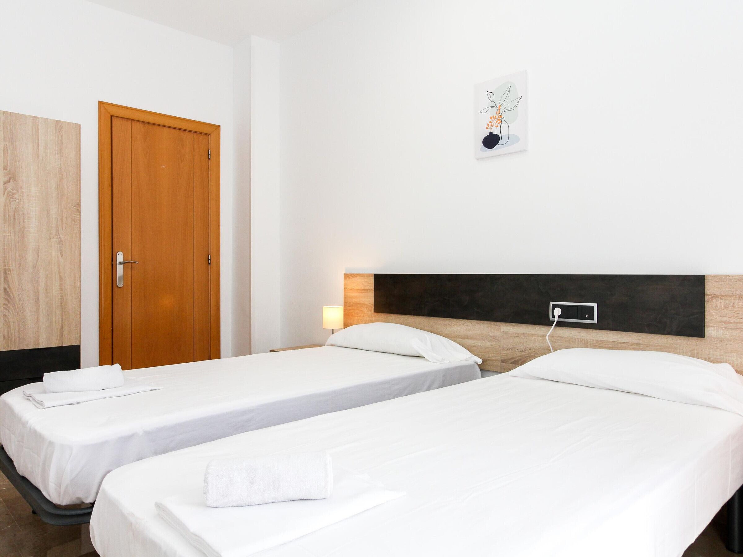 Habitación de hotel con dos camas individuales, una puerta de madera, una lámpara de pared y una pequeña obra de arte.