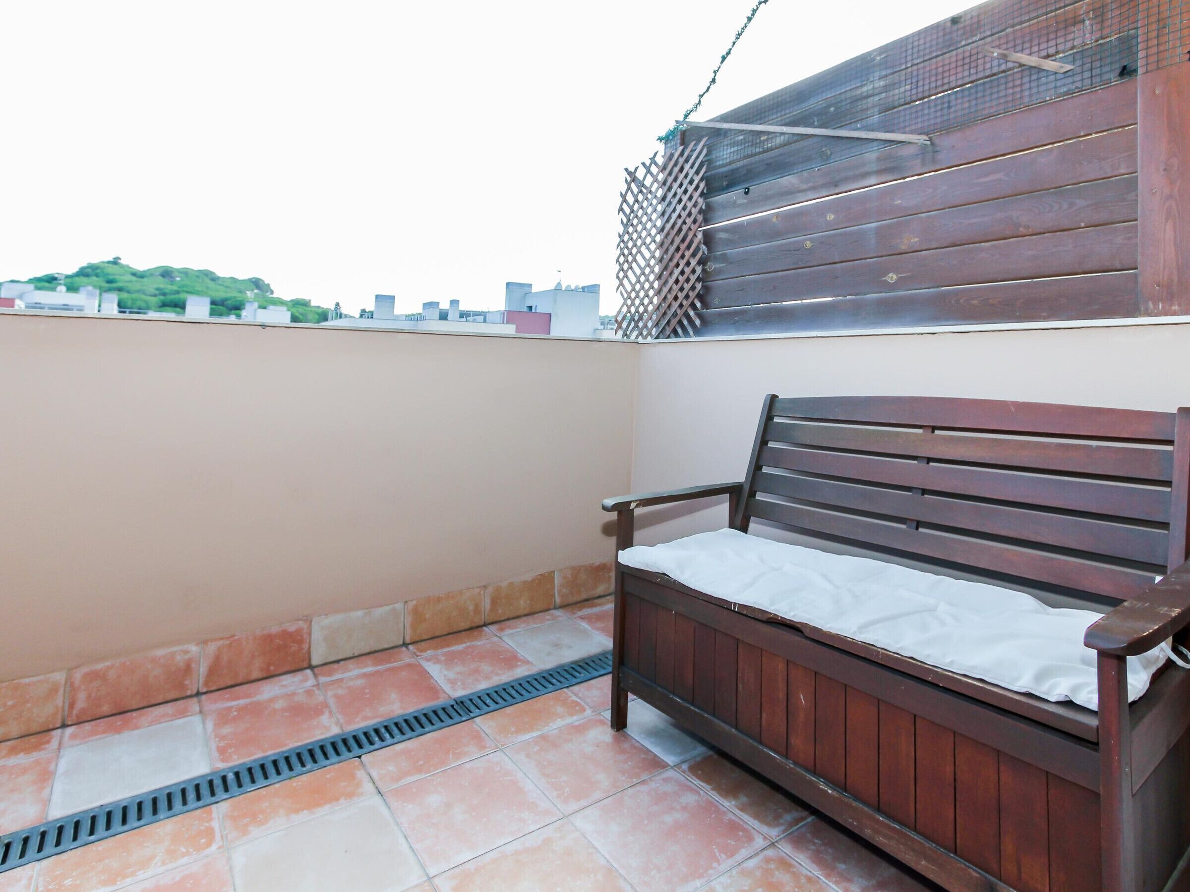 Eine Holzbank mit weißem Kissen auf einem gefliesten Balkon mit einem Metallgitter.