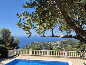 A pool with a stone deck and a white balustrade, overlooking a coastal town and the sea.