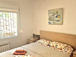 Un dormitorio con cama, una ventana con barrotes, una mesita de noche y un cuadro abstracto colgado en la pared.