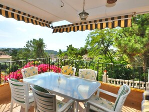A covered outdoor patio with a glass table and white chairs with green cushions. - Modern villa 1 km from beach with private pool and sea views (Lloret de Mar)