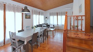 A spacious dining area with a wooden ceiling, white walls, and a tiled floor. There is a dining table with chairs, a sofa, and a staircase with a wooden railing.
