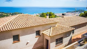 Ein Küstenhaus mit Terrakotta-Dachziegeln, kleinen Fenstern und Blick auf das Meer und die Stadtlandschaft.
