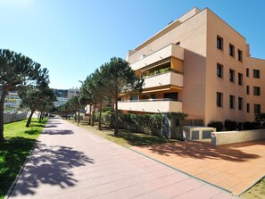 Um edifício moderno de vários andares com varandas, um caminho pavimentado e vegetação ao redor.