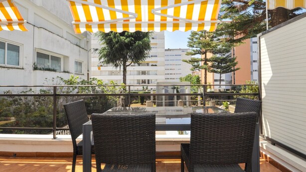 A terrace with two wicker chairs and a glass table, under a striped awning, overlooking a courtyard with buildings and trees. - Cozy New Apartment, Beach 150m, Internet, A/C, Pool Access, Lloret de Mar (Lloret de Mar)