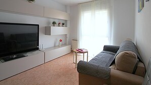 Ein modernes Wohnzimmer mit Flachbildfernseher, einer Couch, einem kleinen Tisch mit Fernbedienung und einem Fenster mit leicht transparenten Vorhängen.