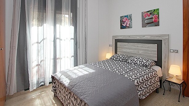 Ein Schlafzimmer mit einem Bett, einem Nachttisch, einer Lampe, einem Fenster mit Vorhängen und zwei Wandbildern.