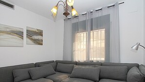 Ein modernes Wohnzimmer mit einem Ecksofa, einem großen Fenster mit durchscheinenden Vorhängen und einer Deckenleuchte.