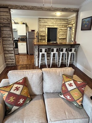 A living room with a sofa and patterned cushions, a kitchen area with white cabinets and a wooden accent wall, and a bar with white stools. - Located 3 miles to Grenada Lake (Grenada)