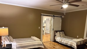 Une chambre avec deux lits, un plancher en bois, un ventilateur de plafond et une porte donnant sur une autre pièce.