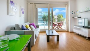A modern living room with a sofa, a dining table, a TV, and a balcony with a view of buildings and greenery. - Charming Family Apartment in Santa Margarita: Pool, Parking, Near Beach and Shops (Roses)