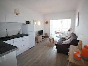 A modern kitchen with white cabinets, a black countertop, and a microwave. There is a dining area with a table and chairs, and a living room with a sofa and a lamp.