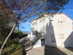 A building with a staircase leading to a balcony, surrounded by greenery and a clear view of the sea.