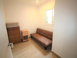 Une petite pièce bien éclairée, avec une commode en bois, une petite table de chevet dotée de tiroirs, un banc et une fenêtre avec des stores.