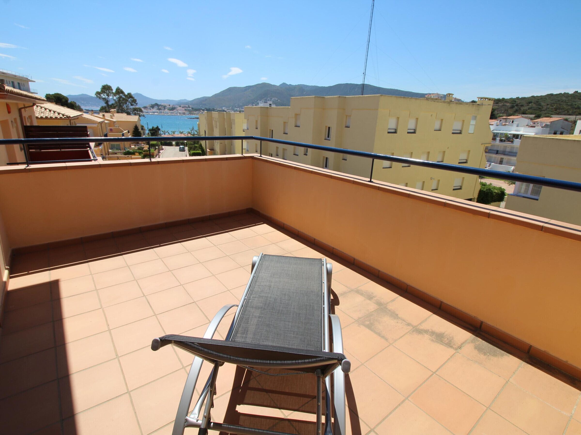 A balcony with a metal chair, tiled floor, and a view of buildings and a body of water in the background.