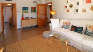 A living room with a white sofa, glass coffee table, and a wooden cabinet. There are framed pictures on the wall and a door leading to another room. - Modern seafront apartment with air conditioning in Llançà (Llançà)