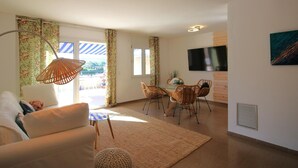 A living room with a sofa, a glass-top coffee table, wicker chairs, a TV on the wall, and a view through the window. - Modern seafront apartment with air conditioning in Llançà (Llançà)