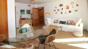 A living room with a glass-top coffee table, wicker chairs, a white sofa, and a dining area with a wooden cabinet and a potted plant. - Modern seafront apartment with air conditioning in Llançà (Llançà)